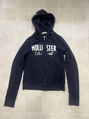 Mörkblå hollister zip-up hoodie - Mörkblå zip-up hoodie från Hollister med broderad vit logga och text på bröstet. Tröjan har huva med snörning, dragkedja framtill och två fickor. Lite urtvättad/nopprig. Vet ej om det är herr/dam modell, storlek M.