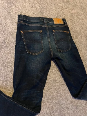 Mörkblå jeans från Nudie Jeans - Säljer ett par mörkblå jeans från Nudie Jeans i storlek 27/32. Jeansen har klassisk femficksdesign, orangea kontrastsömmar och en snygg läderpatch bak i midjan. Materialet är slitstarkt denim och håller riktigt bra. Skriv vid minsta lilla fråga eller fundering‼️