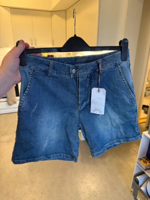 Blå denimshorts från Sira, M - Säljer ett par blå denimshorts från Fira i modellen Fiffi Shorts. De har klassisk femficksdesign, knapp och dragkedja framtill samt bälteshällor. Materialet är slitstark bomullsdenim med lätt slitna detaljer för en chill look. Storlek M. Helt ny med tags/ prislapp fortfarande kvar. 