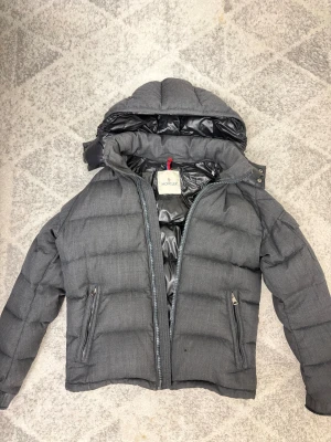  Moncler Montgenevre jacka. 2 - Säljer en mörkgrå pufferjacka från Moncler med huva och snygga detaljer som fickor med dragkedja och logotyp på ärmen.  Perfekt för kalla dagar och malla till sig med. Kvitto medföljs! Nypris på en montgenevre ca 20 000:- säljer billigt pga lite slitage vid ärmänden samt att moncler dragkedjan saknas men går att ersätta och beställa ny dragkedja (den fungerar perfekt redan) Pris kan diskuteras i DM. PRIS KAN DISKUTERAS VID SMIDIG AFFÄR🍾