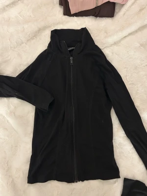 Svart långärmad kofta från Gina Tricot - Svart långärmad kofta från Gina Tricot i storlek XS. Koftan har en hög krage och hel dragkedja framtill, vilket ger en clean och stilren look. Materialet är mjukt och känns bekvämt mot huden, perfekt för lager-på-lager under kyliga dagar.