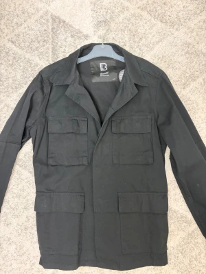 Safarijacka från Brandit - Snygg svart safarijacka från Brandit. Helt och hållet oanvänd och sticker finns kvar i jackan. Stilren och liknar klassiska safarijackorna från Oscar Jacobsson. True to size