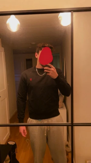 Svart sweatshirt från Ami Paris - Svart sweatshirt från Ami Paris med röd broderad logga på bröstet. Tröjan har rund hals, ribbade muddar vid ärmslut och nederkant samt en clean och stilren look. Tillverkad i mjuk bomull som är skön att bära. Perfekt för dig som gillar minimalistisk design. Perfekt som julklapp