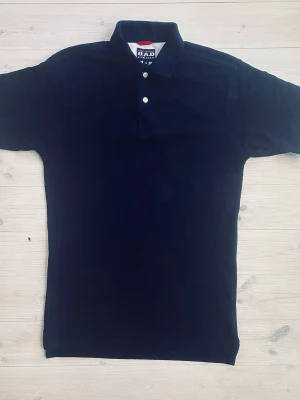 Svart pikétopp från D.A.D Sportswear - Säljer en svart pikétopp från D.A.D Sportswear med klassisk krage och två knappar framtill. Toppen har korta ärmar och är tillverkad i ett slitstarkt material som passar perfekt till sportiga aktiviteter. Stor logga på bröstet i vitt, blått och rött ger en cool touch.