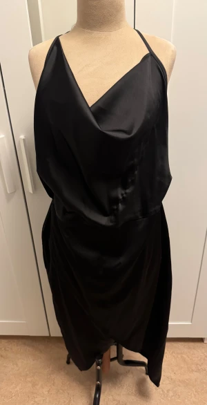 Svart långklänning från Missguided - Snygg svart långklänning från Missguided med tunna axelband och omlott-look framtill. Klänningen har en elegant öppen rygg och är gjord i ett glansigt satinliknande material som ger en lyxig känsla. Perfekt för dig som gillar stilrena och trendiga plagg.
