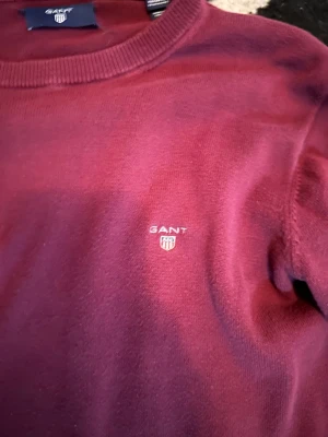 Vinröd stickad tröja från GANT - Snygg vinröd stickad tröja från GANT med rund hals och diskret logga broderad på bröstet. Tröjan har ribbade muddar vid hals, ärmslut och nederkant. Perfekt för dig som gillar klassisk stil med en modern touch. Passar perfekt 155/165