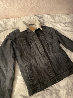 Svart jeansjacka med teddyfoder - Säljer en svart jeansjacka från Levi's med mjukt vitt teddyfoder på insidan och kragen. Jackan har klassisk knappstängning, två bröstfickor med lock och långa ärmar. Perfekt för kyliga dagar och har en riktigt chill vibe.
