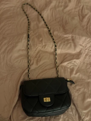 Svart quiltad axelväska från Spanien  - Snygg svart axelväska med quiltat mönster och guldfärgad kedja som axelrem. Väskan har ett klassiskt lock med vridspänne i guld. 