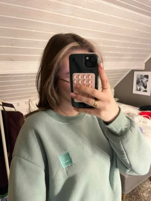 Mintgrön sweatshirt från Acne Studios - Säljer en mintgrön sweatshirt från Acne Studios med deras klassiska ansiktslogga på bröstet. Tröjan har rund hals, ribbade muddar och en loose passform. Perfekt för dig som gillar stilrena och bekväma plagg med lite edge.