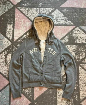 Abercrombie & Fitch cardigan - Säljer en grå hoodie från Abercrombie & Fitch med fluffigt teddyfoder på insidan. Storlek M men är krymp så passar Xs