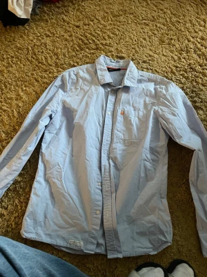 Blåvit randig skjorta Sail Racing - Snygg blåvit randig skjorta från Sail Racing i storlek Large. Skjortan har klassisk krage, bröstficka med orange broderad logga och knappar framtill. Tillverkad i mjuk bomull som är skön mot huden. Perfekt för dig som gillar stilrena detaljer och sportig vibe.