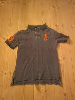 Grå pikétröja från Polo Ralph Lauren - Grå pikétröja från Polo Ralph Lauren med orange broderad logga på bröstet och siffra på ärmen. Klassisk krage och knäppning framtill. Tillverkad i bomull och har korta ärmar. Perfekt för en avslappnad och stilren look.