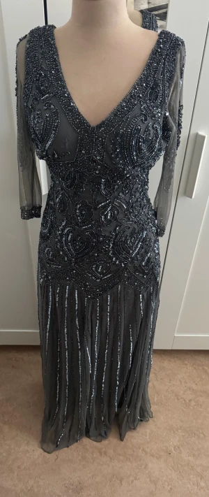 Glittrig långklänning från Piano Nights - Säljer en mörkgrå långklänning från Piano Nights med glittriga paljetter och pärlor, v-ringad fram och bak, samt transparenta långärmar. Klänningen har ett detaljerat mönster och är tillverkad i rayon med polyesterfoder. Perfekt för dig som vill sticka ut.