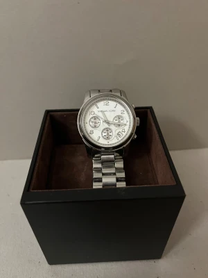 MK Klocka - Bilderna visar en silver/vit klocka från Michael Kors med länkarmband och extra länkar. Orginalförpackning med brun mocha för att minska skrapor på klockan. Använd men i bra skick. Batteriet byttes senast för 1 år sedan. Om du har frågor är det bara att höra av dig.