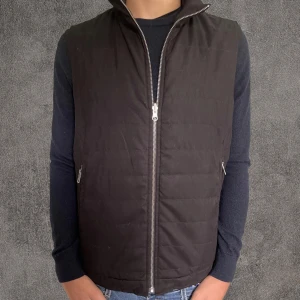  J.Lindeberg Manchester väst - Stilren svart Manchester väst från J.Lindeberg med hög krage och hel dragkedja framtill. Västen har två fickor med dragkedja och quiltad design. Materialet är en mix av ull och polyamid som ger en mjuk men stadig känsla. Perfekt att slänga över en hoodie eller t-shirt.