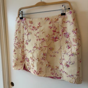 Beige blommig kortkjol med broderi - Superfin beige kortkjol med broderade blommor i rosa, lila och gult. Kjolen har en rak passform och är fodrad med rosa tyg på insidan. Endast testad aldrig använd. 