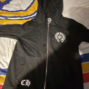 Svart Chrome Hearts hoodie med tryck - Svart hoodie från Chrome Hearts med stora vita tryck på både fram- och baksida samt ärmar. Dragkedja framtill, huva med snörning och två fickor. Snygg streetwear-vibe med Los Angeles-tryck och ikoniska CH-loggor. Perfekt för dig som gillar bold designs.