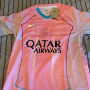 PSG x Nike rosa fotbollströja S - Säljer en PSG fotbollströja från Nike i rosa och ljusblå toner. Tröjan har korta ärmar, V-ringning med blå detalj och tryck med Qatar Airways på bröstet. Materialet är lätt och andas, perfekt för match eller träning. Snyggt PSG-emblem och Nike-logga på framsidan.