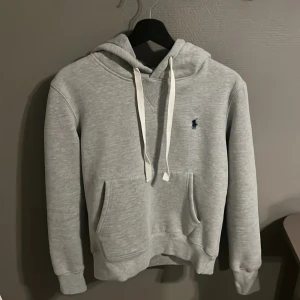 Grå hoodie från Polo Ralph Lauren - Snygg ljusgrå hoodie från Polo Ralph Lauren med klassisk broderad logga på bröstet. Tröjan har huva med vita snören, känguruficka och mjukt material på insidan. Perfekt för chill dagar och enkel att matcha med jeans eller joggers.