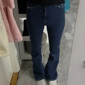 Blå bootcut jeans med hög midja - Snygga blå jeans med bootcut passform och hög midja. Klassisk femficksmodell i jeansmaterial, perfekt för dig som gillar en tidlös look med lite utsvängda ben. Färgen är mörkblå och byxorna har en rak siluett från höften och nedåt.