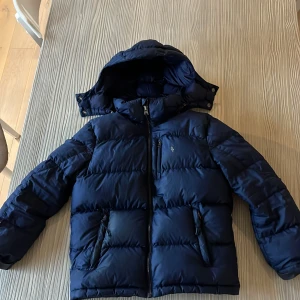 Mörkblå pufferjacka Polo Ralph Lauren - Snygg mörkblå pufferjacka från Polo Ralph Lauren med huva och dragkedja framtill. Jackan har två fickor med dragkedja och klassisk Polo-logga på bröstet. Perfekt för kalla dagar och riktigt skön att bära. Den har en skada på ryggenen syns inte på långt håll. Pågrund av den lilla skadan så är priset jättebra. Men pris kan diskuteras.