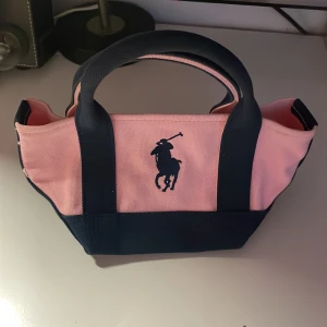 Rosa och marinblå handväska från Polo Ralph Lauren - Snygg liten handväska från Polo Ralph Lauren i rosa och marinblå bomullscanvas. Väskan har två rejäla handtag och klassisk broderad polospelare på framsidan. Perfekt storlek för det viktigaste och ger en sportig men söt vibe.