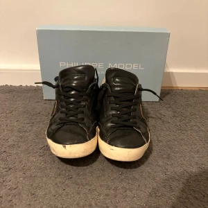 Svarta Philippe Model sneakers - Snygga svarta sneakers från Philippe Model med vita sulor och silverdetalj på hälen. Klassisk låg modell med svarta skosnören och ikonisk sköld på sidan. Perfekta för dig som gillar stilrena och trendiga skor.