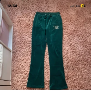 Gröna velourbyxor Juicy Couture - Mörkgröna mjukisbyxor från Juicy Couture i mjuk velour med bootcut-ben. Byxorna har snörning i midjan, två bakfickor och ett glittrigt JC-logo i strass på framsidan. Perfekta för en chill och trendig look.