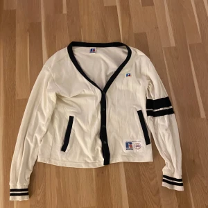 Vit cardigan från Russell Athletic - Snygg vit kofta från Russell Athletic med svarta detaljer och knappar framtill. V-ringad med svarta ränder på ena ärmen och muddar. Två fickor och patch med logga nedtill. Perfekt för dig som gillar sportig och retro stil.
