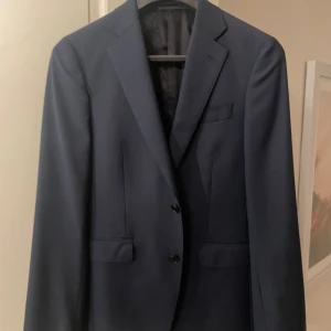 Marinblå kavaj These Glory Days - Snygg marinblå kavaj från These Glory Days med klassisk tvåknäppning och smal passform. Kavajen har fickor med lock och en bröstficka, samt slät baksida. Perfekt för dig som gillar stilrena och tidlösa plagg.