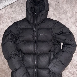 Svart Peak Performance dunjacka - Svart dunjacka från Peak Performance Rivel down jacket, med huva och diskret logga på bröstet och ärmen. Jackan har quiltad design, dragkedja framtill och två sidofickor. Perfekt för kalla dagar och har en clean, modern look som funkar till streetwear.  Se länk till jacka i butik: https://www.peakperformance.com/se/sv/product/m-rivel-jacket-g77962.html.                                 Axel t axel ca 45. Axel t botten av jackan ca 65. Min son är 192cm lång och smal, han kan nästan ha den.