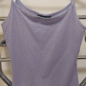 Glittrig lila linnetopp med smala band - Säljer en snygg lila linnetopp från Att i storlek M. Toppen har smala axelband och är glittrig med en skimrande yta som syns tydligt i ljuset. Perfekt för dig som vill sticka ut med en cool och trendig look. Materialet är mjukt och följsamt.