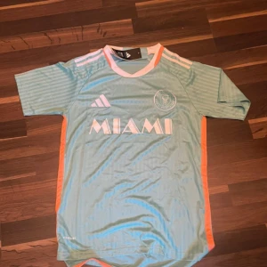 Inter Miami FC ljusblå matchtröja Adidas - Snygg ljusblå Inter Miami FC fotbollströja från Adidas med korta ärmar och diskreta ränder. Tröjan har rosa detaljer på axlar och sidor, samt klubbmärke och MIAMI-tryck på bröstet. Tillverkad i lätt och ventilerande polyester med AEROREADY-teknologi.