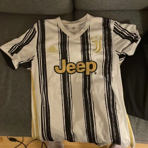 Juventus fotbollströja Adidas M - Juventus matchtröja från Adidas i storlek M. Tröjan är vit med svarta ränder och har guldiga detaljer som Jeep-loggan, Adidas-märket och Juventus-emblemet med tre stjärnor. Tillverkad i lätt och ventilerande polyester, perfekt för fotboll eller häng.
