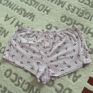 Rosa shorts med körsbärsmönster - Säljer ett par ljusrosa shorts med gulligt körsbärsmönster och knappar framtill. De har en loose passform och är gjorda i mjuk bomull, perfekta för chill dagar hemma eller som pyjamasshorts.