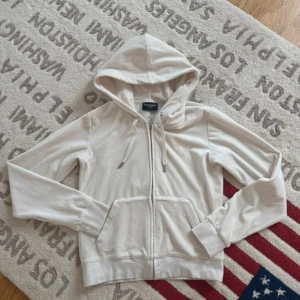 Beige velour hoodie från Juicy Couture - Supermjuk beige hoodie från Juicy Couture i velour med dragkedja och huva. Tröjan har snörning i halsen, broderad JC-logga på bröstet och snygg J-detalj på dragkedjan. Perfekt för en chill och trendig look.