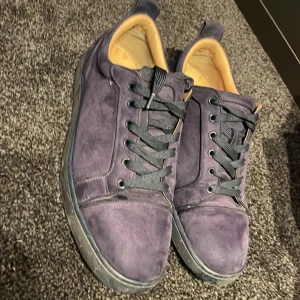 Blå mockasneakers från Louboutin - Säljer ett par snygga sneakers från Christian Louboutin i mörkblå mocka med svarta skosnören och ikoniska röda sulor. Skorna har rund tå, platt sula och broderad Louboutin-logga på hälen. Insidan är beige och de har klassisk snörning framtill.