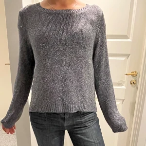 Glittrig stickad tröja - Säljer en mörkgrå glittrig stickad tröja med rundad hals och långa ärmar. Tröjan har en enkel, klassisk design med ribbade muddar vid ärmslut och nederkant. Perfekt för dig som gillar en chill och stilren look.