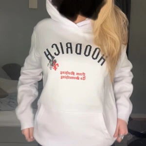 Vit hoodie med tryck från Hoodrich - Säljer en vit hoodie från Hoodrich med svart och rött tryck framtill och coola detaljer. Tröjan har huva, magficka och långa ärmar. Perfekt för dig som gillar streetwear och vill ha en chill look.