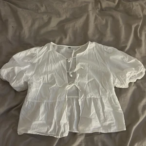 Vit blus med puffärm och knyt - Säljer en söt vit blus med puffiga korta ärmar och tre knytband framtill. Blusen har en luftig och flowy passform med volang nedtill. Perfekt för dig som gillar romantisk och chill stil. Materialet känns som bomull och är lätt och skönt.