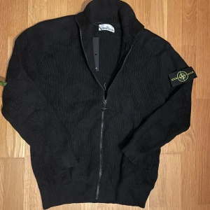 Svart stickad kofta  - Svart stickad kofta med ribbad struktur och hel dragkedja framtill. Klassisk logotyp-patch på ärmen och snyggt detaljerad dragkedja med Stone Island-emblem. Perfekt för dig som gillar streetwear och vill ha något stilrent och bekvämt.