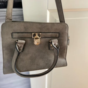 Grå mocka handväska med gulddetalj - Snygg grå handväska i mocka med stilrena svarta kanter och två handtag. Väskan har en dekorativ guldfärgad låsdetalj framtill och praktisk dragkedja på sidan. Perfekt storlek för att få plats med allt viktigt.