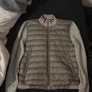 Grå Moncler Down Cardigan - Heatnlux - Säljer en grå dunväst från Moncler med quiltad framsida och mjuka stickade ärmar. Västen har dragkedja, ribbad krage med röd och vit rand, samt Moncler-logga på ärmen. Perfekt för lager-på-lager och snygg streetstil. Moncler loggan på insidan där man hänger den har tråden åkt av men det går lätt att fixa👍🏽pris går att diskuteras privat👌🏽