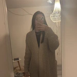 Beige stickad kofta med bälte - Säljer en chunky beige stickad kofta med breda ribbade muddar och tillhörande bälte i midjan. Koftan har lång ärm och en loose, oversized passform som ger en chill vibe. Perfekt att slänga på sig när det är lite kallare ute.