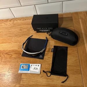 Oakley radar ev path - Säljer ett par Oakley sportglasögon med blå och silverfärgade bågar och svarta linser. Modellen har en modern, halvbåge-design och kommer med originalfodral, putsduk och mjuk påse. Perfekt för dig som vill ha en cool och funktionell look när du är aktiv.