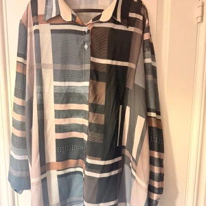 Oversized skjorta med grafiskt mönster - Snygg oversized skjorta med grafiskt rutigt mönster i svart, beige, grått och brunt. Skjortan har långa ärmar, klassisk krage och knappar framtill. Perfekt för dig som gillar statement-plagg med coola färgkombinationer och unik design.