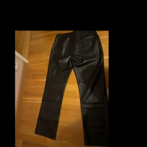 Svarta raka byxor i skinnimitation - Säljer ett par svarta byxor i skinnimitation med raka ben och klassisk femficksdesign. Snyggt stilrena och passar perfekt till en cool streetstil. Materialet är syntetiskt och ger en lyxig känsla. Byxorna har normal passform och midwaist.