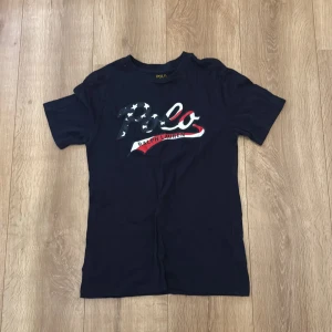 Mörkblå Polo Ralph Lauren t-shirt - Snygg mörkblå t-shirt från Polo Ralph Lauren med stort Polo-tryck på bröstet i amerikanska flaggans färger. Klassisk rund hals och korta ärmar. Tillverkad i mjuk bomull som känns skön mot huden. Perfekt för dig som gillar streetwear och ikoniska märken.