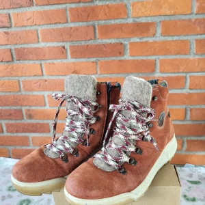 Rieker bruna mockaboots med ullfoder - Snygga bruna boots från Rieker i mocka med grov sula och färgglad stickad kant upptill. Skorna har ullfoder, dragkedja på sidan och mönstrade snören i rött och vitt. Perfekta för höst och vinter med sin varma insida och coola outdoor-vibe.