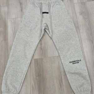 Grå mjukisbyxor Essentials Fear of God - Snygga grå mjukisbyxor från Essentials Fear of God med elastisk midja och snörning. Byxorna har en relaxed passform, ribbade muddar vid bensluten och tryckt logga på benet. Perfekta för chill eller streetwear. Materialet är mjukt och skönt bomullsmix. KOMMER MED ORIGINAL ESSENTIALS ZIP BAG.